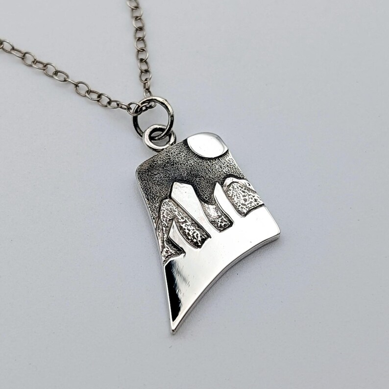 May include: A silver-toned pendant necklace featuring a trapezoidal charm with a textured, dark-finished mountain range and a circular moon design. The pendant hangs from a delicate silver-toned chain with round links, showcasing a polished finish.