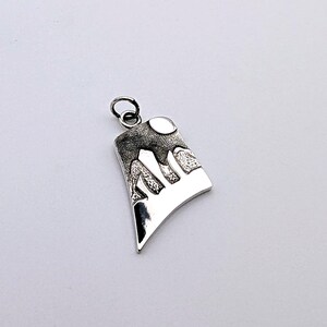 May include: A silver-toned pendant featuring a stylized mountain landscape with a textured, dark upper section and a smooth, polished lower section. The design includes a circular moon shape at the top and a small jump ring attached for hanging.