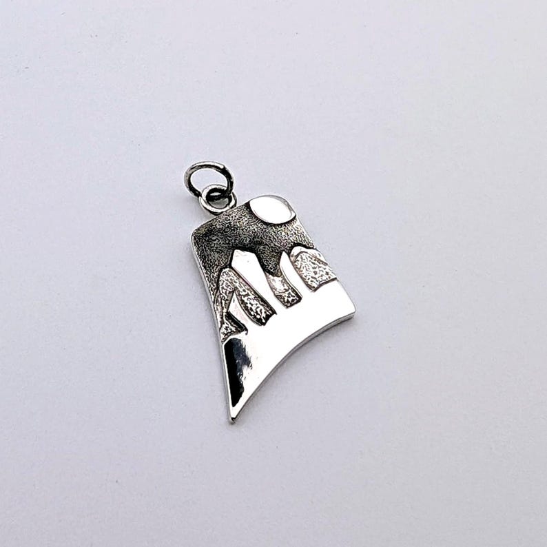 May include: A silver-toned pendant featuring a stylized mountain landscape with a textured, dark upper section and a smooth, polished lower section. The design includes a circular moon shape at the top and a small jump ring attached for hanging.