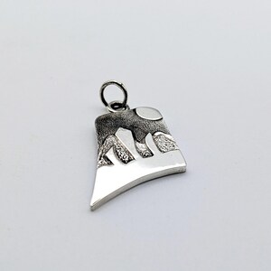 May include: A silver-toned metal pendant featuring a stylized landscape design with a textured dark upper section depicting mountains and a circular moon, contrasted against a smooth, polished lower section. The piece includes a small jump ring.