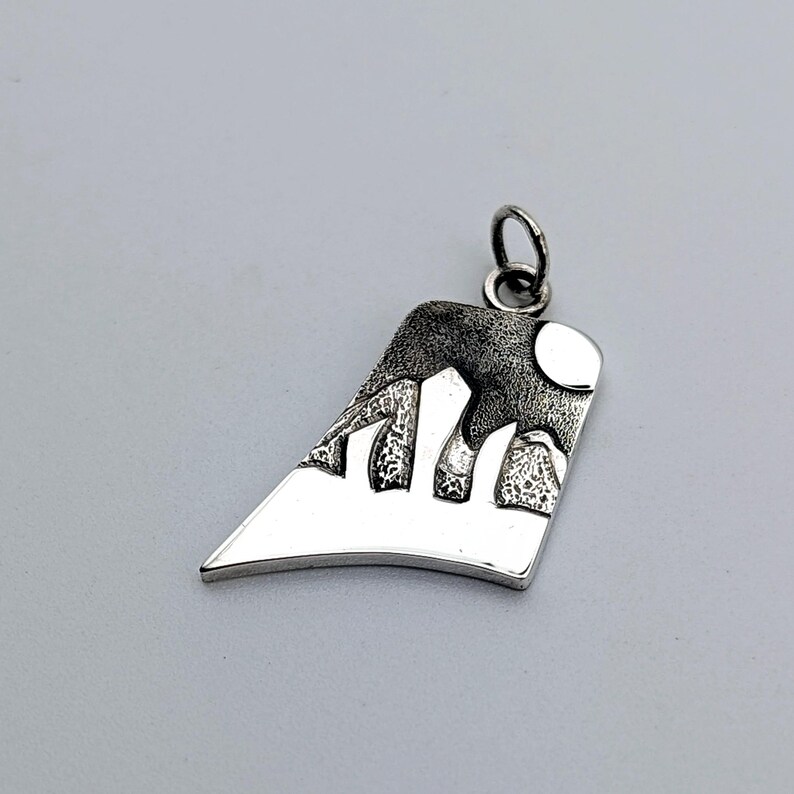 May include: A silver-toned pendant with an asymmetrical, trapezoidal shape. The surface features a textured, dark-oxidized mountain range design against a smooth, polished silver base, topped with a small circular jump ring for attachment.