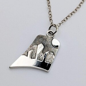 May include: A silver pendant necklace featuring an asymmetrical, trapezoidal shape with a textured, dark oxidized finish depicting a mountain range and a circular moon. The pendant hangs from a delicate silver chain with round links.