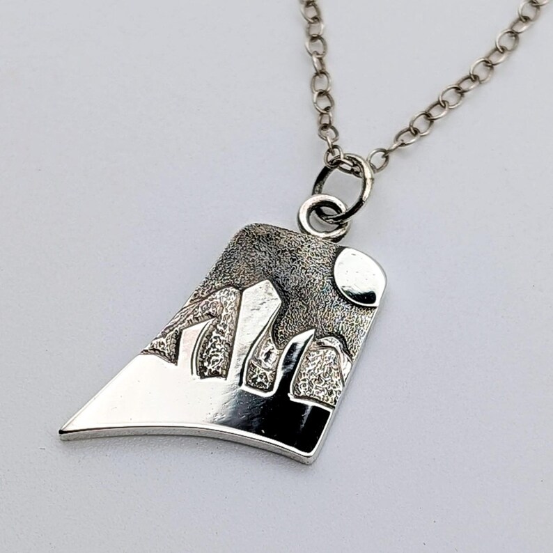 May include: A silver pendant necklace featuring an asymmetrical, trapezoidal shape with a textured, dark oxidized finish depicting a mountain range and a circular moon. The pendant hangs from a delicate silver chain with round links.