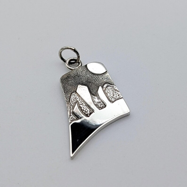 May include: A silver pendant featuring a stylized mountain landscape with a textured dark sky and a smooth, polished moon. The design includes sharp, angular peaks and a solid black base, finished with a small circular jump ring at the top.