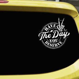 May include: A white circular vinyl decal applied to a car window featuring a skeleton hand making a peace sign. The text reads HAVE THE DAY YOU DESERVE in a stylized font, arranged within a circular border with small star and lightning accents.