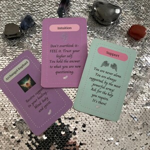 May include: Three purple, green, and blue oracle cards with angel wings and inspirational messages. The cards read "Archangel Raphael", "Intuition", and "Support".