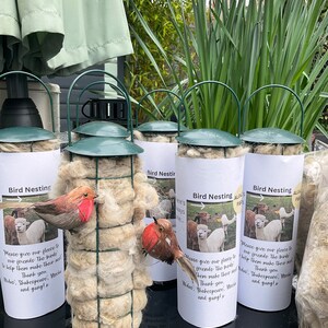 May include: Five white cylindrical containers with green lids and wire cages filled with white fluffy material. Each container has a white label with the text "Bird Nesting" and a picture of a bird or alpaca. The text on the label reads "Please give our fleece to our friends. The birds need them make their nests. Thank you. Wdae: Shakespeare, Mocha and gang! x"