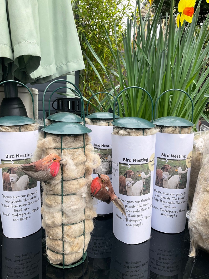 May include: Five white cylindrical containers with green lids and wire cages filled with white fluffy material. Each container has a white label with the text "Bird Nesting" and a picture of a bird or alpaca. The text on the label reads "Please give our fleece to our friends. The birds need them make their nests. Thank you. Wdae: Shakespeare, Mocha and gang! x"