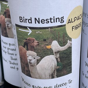 May include: A white cup with a brown and white alpaca fleece on it. The cup has a yellow circle with the text "Alpaca Fibre" on it. The text "Bird Nesting" is at the top of the cup. The text "Please give our fleece to our friends, the birds, to help them make their nest." is at the bottom of the cup.