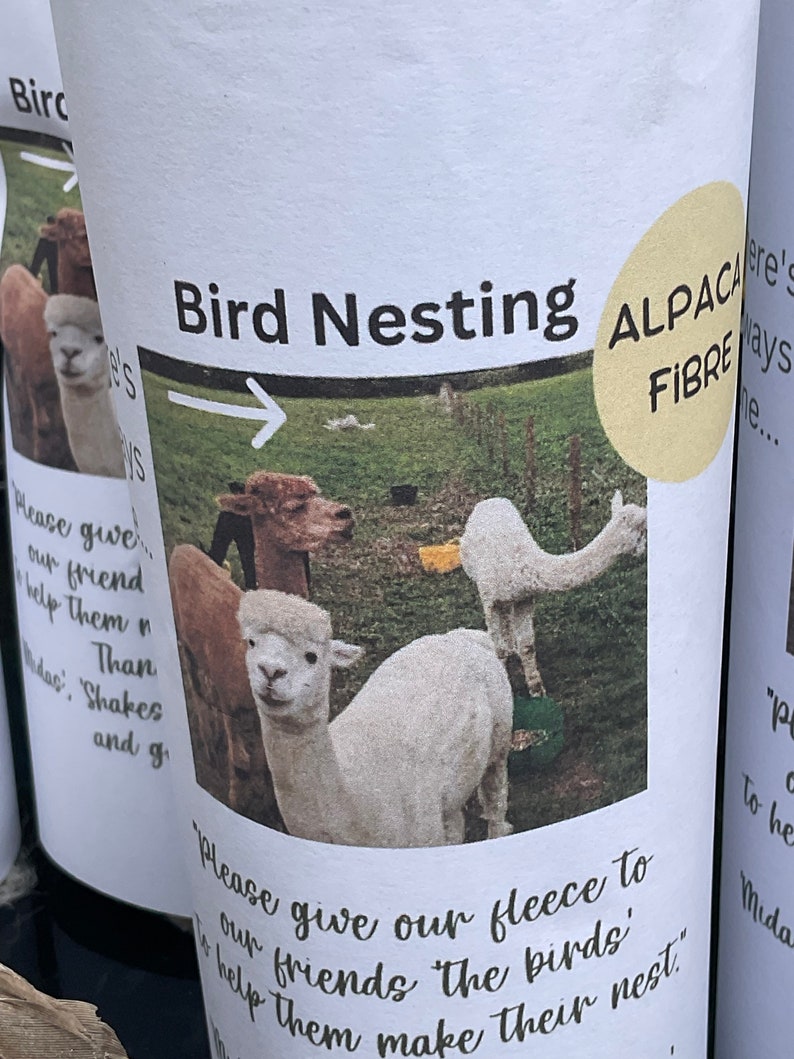 May include: A white cup with a brown and white alpaca fleece on it. The cup has a yellow circle with the text "Alpaca Fibre" on it. The text "Bird Nesting" is at the top of the cup. The text "Please give our fleece to our friends, the birds, to help them make their nest." is at the bottom of the cup.