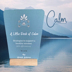 May include: A blue card with a white floral design and the text "A Little Deck of Calm" and "Strategies to support a healthy mindset. (With Affirmations) 36 cards By Amanda Anderson". The card is standing on a wooden block against a background of a lake and mountains. The word "Calm" is written in blue cursive above the card.