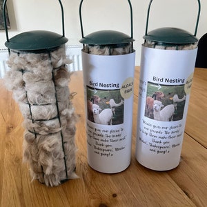 May include: Three green bird feeders filled with alpaca fleece. The feeders have a white label with the text "Bird Nesting Alpaca Fibre" and a message from the alpaca farm owners thanking customers for helping birds make their nests.