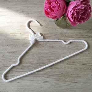 May include: A white padded clothes hanger with a small bow, set against a light wood grain surface. A small glass vase holds three vibrant pink peonies and green leaves, adding a touch of color to the scene.