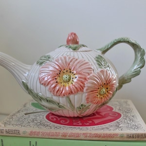 Puede incluir: Una tetera de cerámica blanca con un diseño floral. La tetera tiene un cuerpo redondo con un diseño en relieve de flores rosas y amarillas. La tetera tiene un asa y un pico verdes.