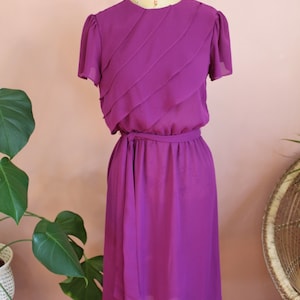 May include: A purple midi dress with a draped bodice and a self-tie belt. The dress has short sleeves and a round neckline.