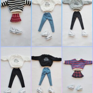 May include: Six different doll outfits with a variety of tops, bottoms, and skirts. The outfits include a striped sweater, a white sweater with a graphic, a gray sweater with a graphic, a black sweater with a graphic, a white sweater with a graphic, and a striped sweater. The bottoms include black leggings, blue jeans, and a plaid skirt. The outfits are all paired with white sneakers.