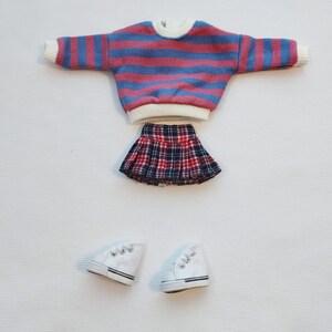 May include: Set of doll clothes, including a striped sweater, plaid skirt, and white sneakers. The sweater is pink and blue with white trim. The skirt is red, black, and white plaid. The sneakers are white with black trim.