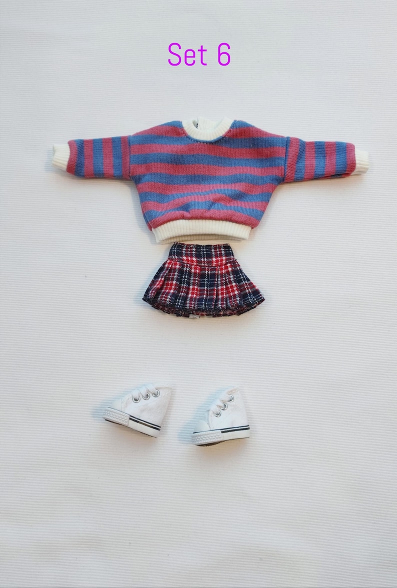May include: Set of doll clothes, including a striped sweater, plaid skirt, and white sneakers. The sweater is pink and blue with white trim. The skirt is red, black, and white plaid. The sneakers are white with black trim.