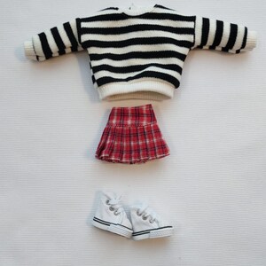 May include: Set 2 of doll clothes includes a black and white striped sweater, a red and black plaid skirt, and a pair of white sneakers.