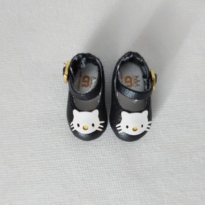May include: A pair of black Mary Jane style shoes with a white Hello Kitty face on the toe. The shoes have a gold buckle and a strap. The shoes are small, likely for a child or doll.