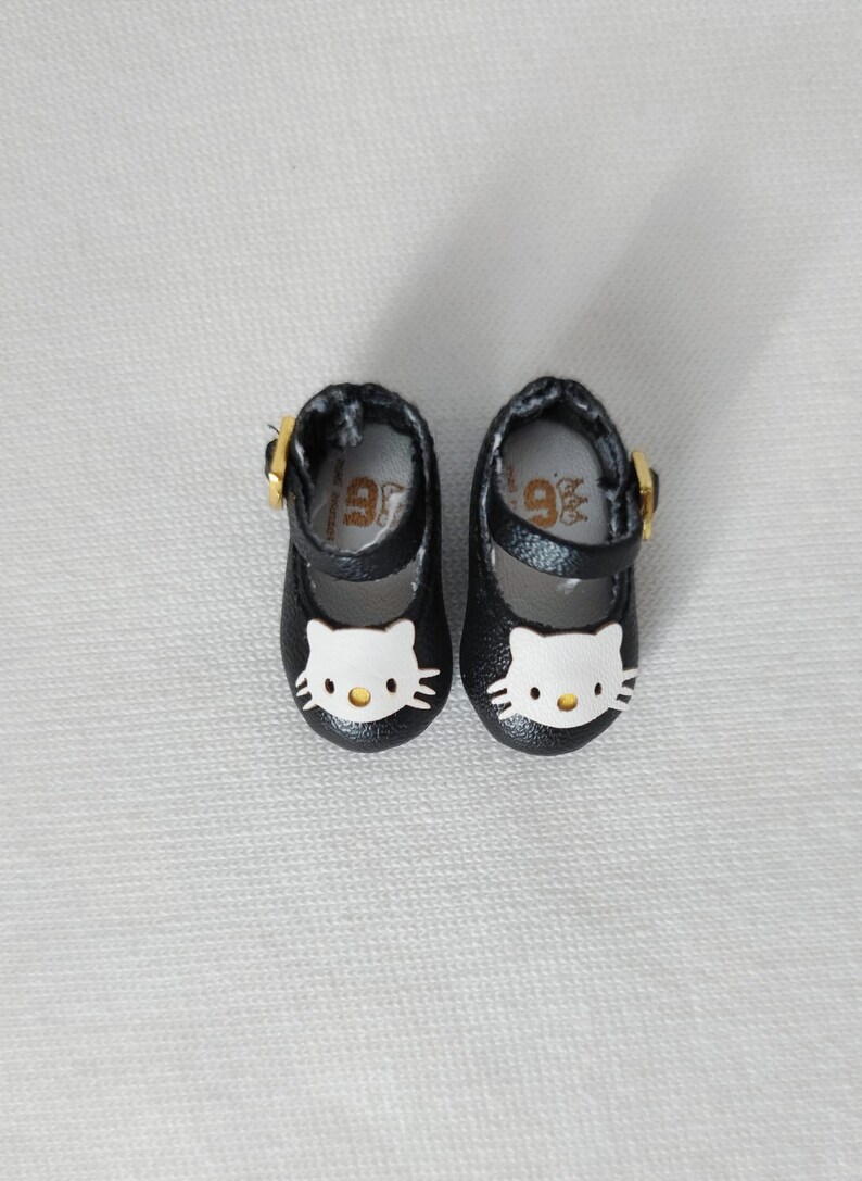 May include: A pair of black Mary Jane style shoes with a white Hello Kitty face on the toe. The shoes have a gold buckle and a strap. The shoes are small, likely for a child or doll.