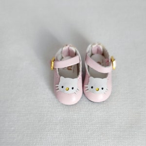 May include: A pair of pink doll shoes with a white cat face design on the toe. The shoes have a strap with a gold buckle. The shoes are made of a soft material and are designed for a doll.