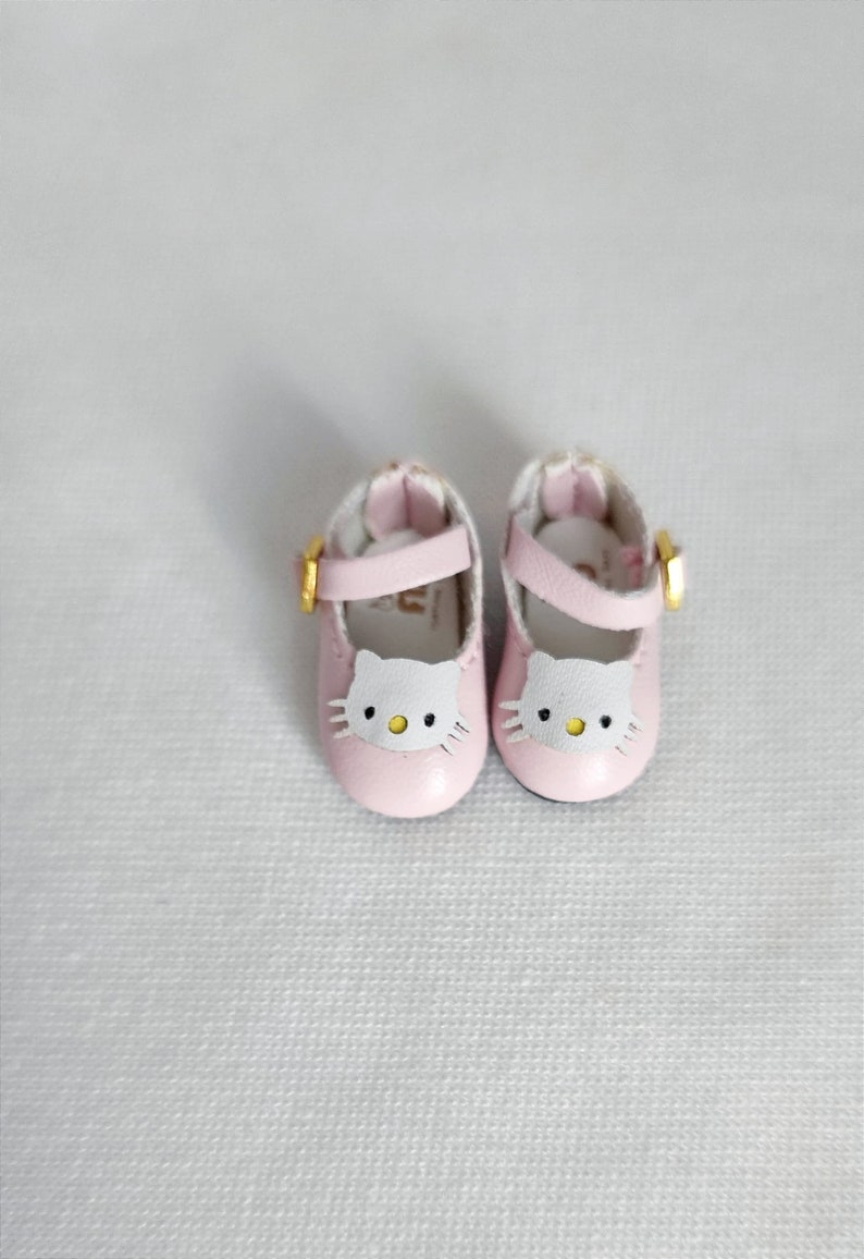 May include: A pair of pink doll shoes with a white cat face design on the toe. The shoes have a strap with a gold buckle. The shoes are made of a soft material and are designed for a doll.