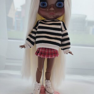 May include: A doll with long blonde hair, wearing a black and white striped sweater, a red plaid skirt, and white sneakers. The doll is standing on a pink stand.