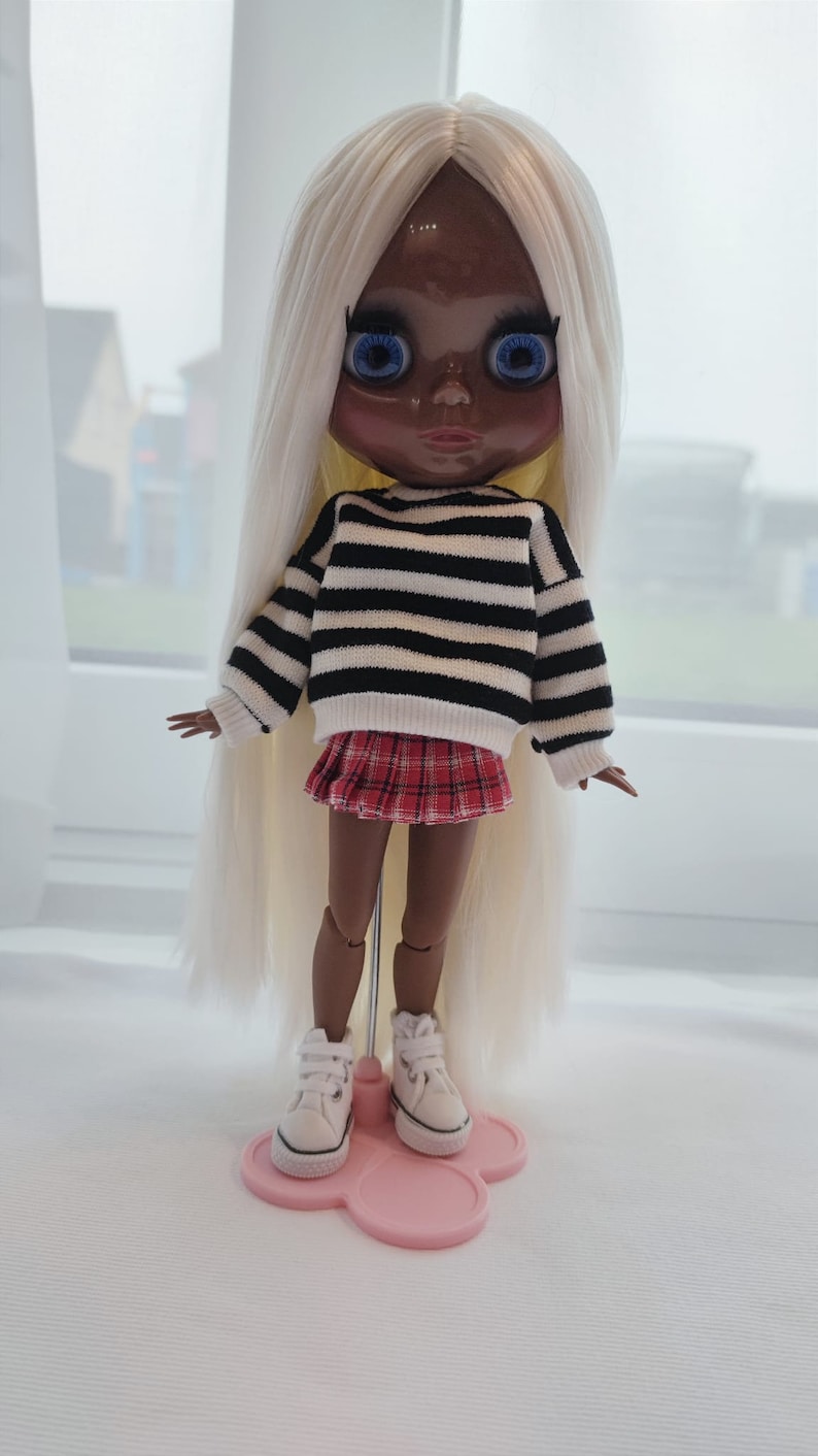 May include: A doll with long blonde hair, wearing a black and white striped sweater, a red plaid skirt, and white sneakers. The doll is standing on a pink stand.