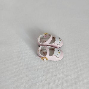 May include: A pair of light pink Mary Jane style shoes with a white cat face design on the toe. The shoes have a small gold buckle and a strap. The shoes are designed for a doll or small child.