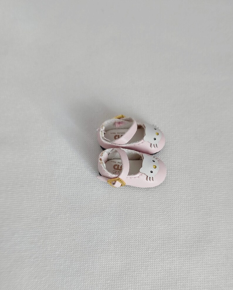 May include: A pair of light pink Mary Jane style shoes with a white cat face design on the toe. The shoes have a small gold buckle and a strap. The shoes are designed for a doll or small child.