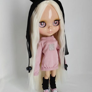 Könnte beinhalten: Eine Puppe mit langen blonden Haaren, die einen rosa Pullover mit Strassstein-Design, schwarze Leggings und weiße Turnschuhe trägt. Die Puppe trägt eine schwarze Kapuze mit Bärenohren.