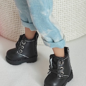 Może przedstawiać: Para miniaturowych czarnych butów z białymi przeszyciami i srebrnymi oczkami. Buty są sznurowane i mają grubą podeszwę. Buty są noszone z jasnoniebieskimi i białymi spodniami.