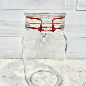 May include: A clear glass jar with a red wire closure. The jar has a textured surface and is empty.