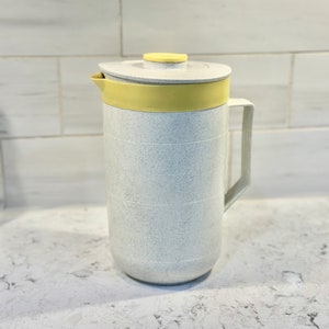 May include: A white pitcher with a speckled finish and a yellow lid and handle. The pitcher is sitting on a white countertop.
