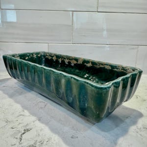 May include: A rectangular, teal-colored ceramic planter with a ribbed design. The planter has a glossy finish and is suitable for indoor or outdoor use. The planter's dimensions are approximately 12 inches long, 5 inches wide, and 4 inches tall.