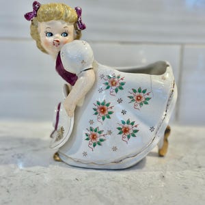 May include: A vintage porcelain planter shaped like a girl wearing a white dress with pink and green floral accents. She has blonde hair with purple bows and is holding a hat.