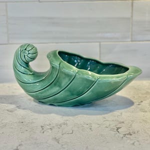 May include: A green ceramic bowl shaped like a seashell with a curled handle. The bowl has a smooth, glossy finish and is perfect for serving snacks or holding small items.