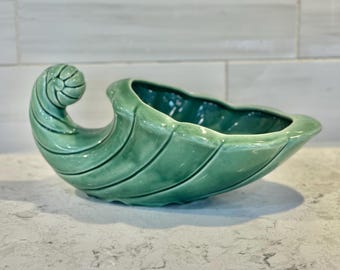 May include: A green ceramic bowl shaped like a seashell with a curled handle. The bowl has a smooth, glossy finish and is perfect for serving snacks or holding small items.