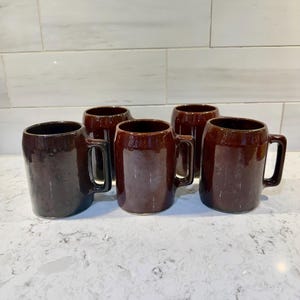 May include: Five brown ceramic mugs with handles are arranged on a white countertop. The mugs have a dark, glossy finish and a cylindrical shape. The mugs are of similar size and design, suggesting they are part of a set.