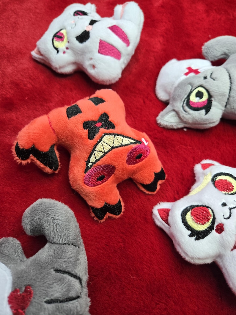 May include: A collection of plush toys in various shapes and colors, including orange, white, and gray. The toys have embroidered details, such as teeth, eyes, and markings. They are arranged on a red surface.