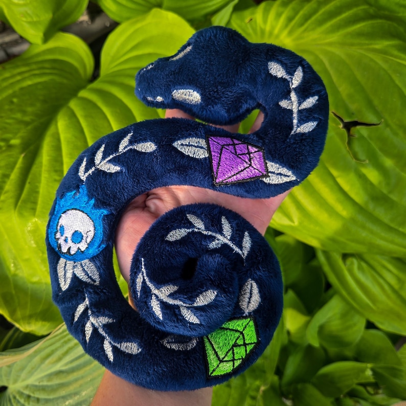 Op de afbeelding: Een donkerblauw pluche slangenknuffel met geborduurde details. De slang heeft een schedel met blauwe vlammen, geometrische paarse en groene edelstenen en zilveren bladaccenten. Het speelgoed wordt in een hand gehouden.