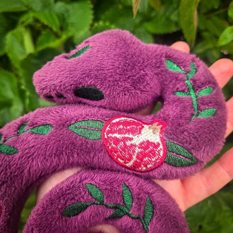 May include: Plush purple snake toy with green embroidered leaves and a red and white pomegranate design. The snake is made of soft, fuzzy material and is held in a hand against a background of green leaves.