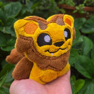 May include: A plush lion toy with a yellow body, brown mane, and orange accents. The lion has embroidered black eyes and a smiling mouth. The toy is made of soft, fuzzy material and is held in a hand against a background of green leaves.