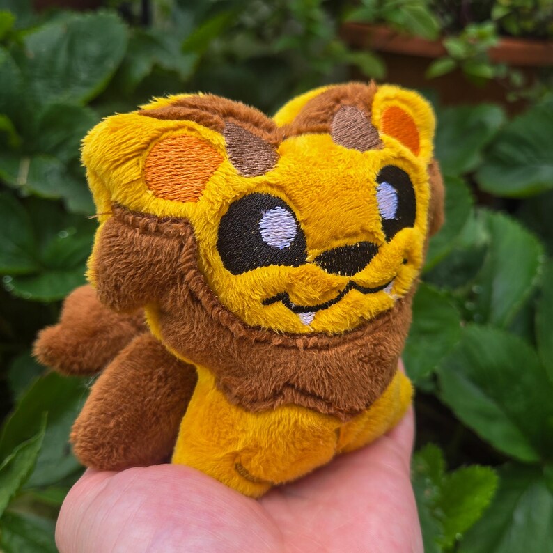 May include: A plush lion toy with a yellow body, brown mane, and orange accents. The lion has embroidered black eyes and a smiling mouth. The toy is made of soft, fuzzy material and is held in a hand against a background of green leaves.