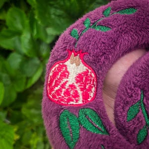 May include: Close-up of a plush, purple item with an embroidered pomegranate design. The pomegranate is red and white, with green leaves and stems. The item has a soft, textured appearance.