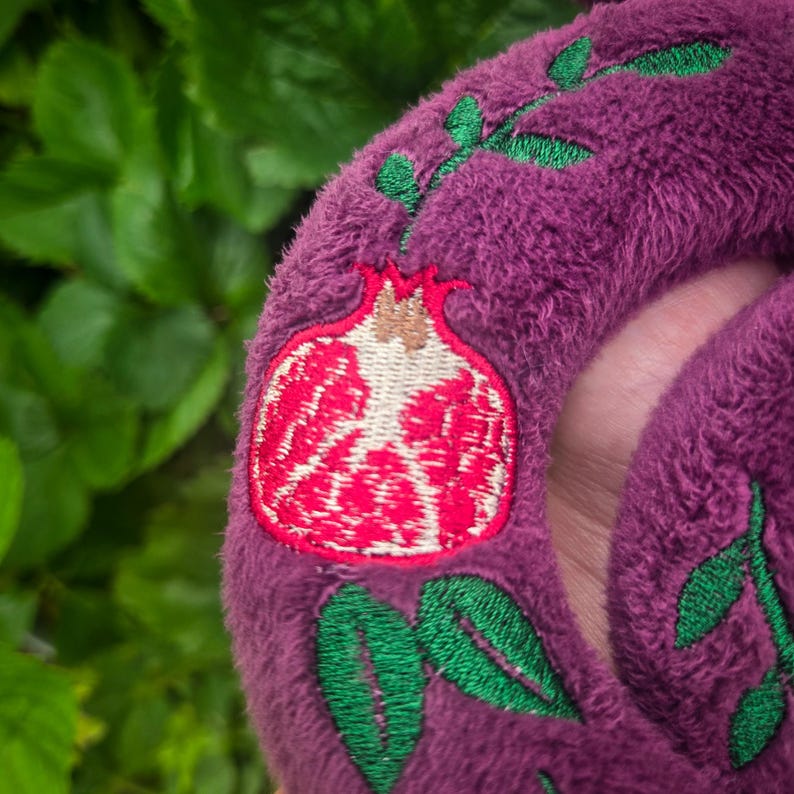 May include: Close-up of a plush, purple item with an embroidered pomegranate design. The pomegranate is red and white, with green leaves and stems. The item has a soft, textured appearance.