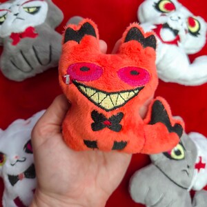 May include: A collection of plush cat toys in various colors. The central toy is orange with black accents, a stitched smile, and pink eyes. Other cats are white and gray, some with bowties and medical symbols.
