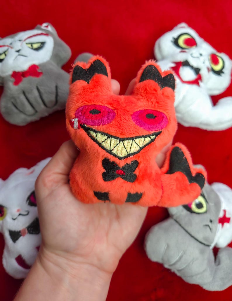 May include: A collection of plush cat toys in various colors. The central toy is orange with black accents, a stitched smile, and pink eyes. Other cats are white and gray, some with bowties and medical symbols.