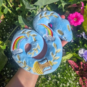 May include: A light blue plush snake toy featuring embroidered gold leaf vines, two rainbows with grey clouds, and a gold pitcher. The soft fabric is shaped into a coiled form, showcasing detailed stitching and vibrant colors on the surface.